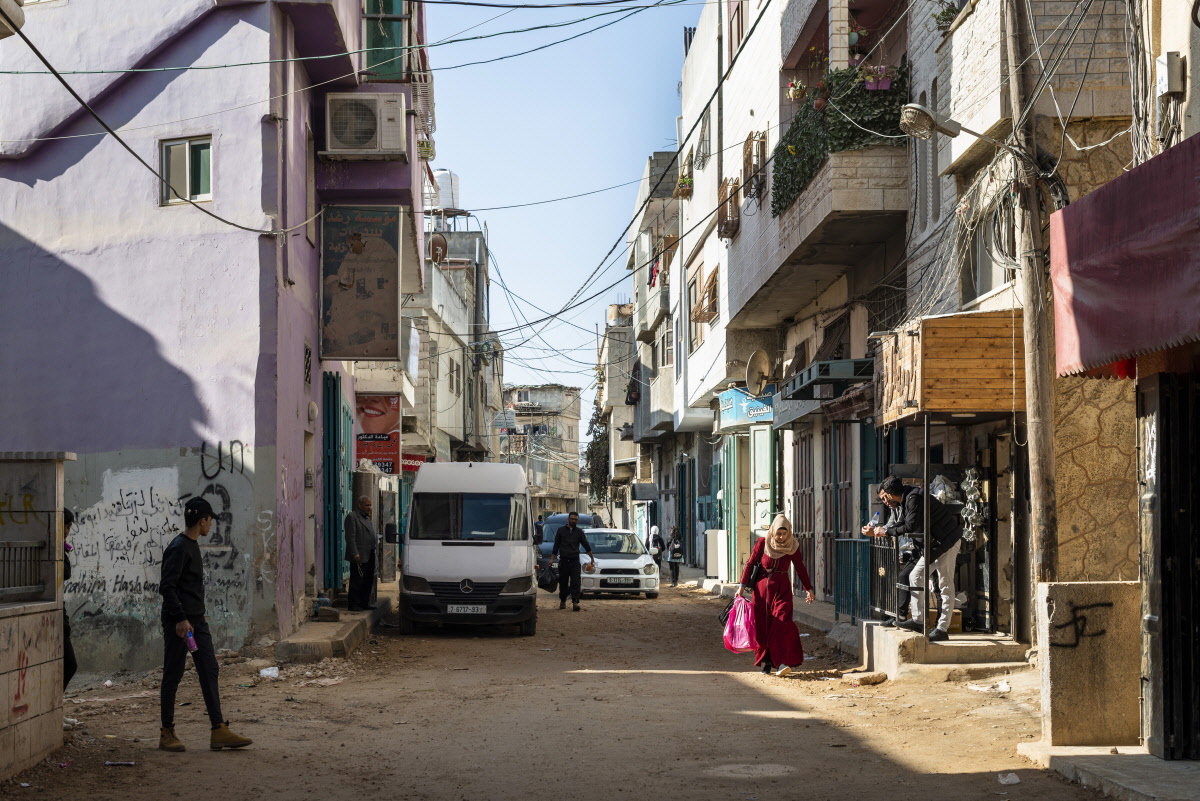 A sunlit street in Arroub Refugee Camp, Palestine, with a woman in a long red dress walking past shopfronts