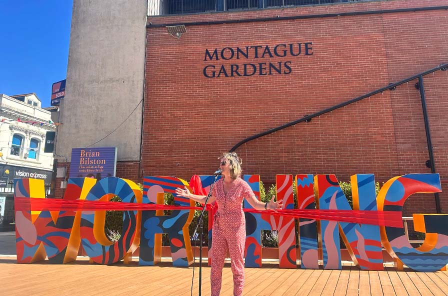 Woman in pink outfit speaking into microphone on decking with colourful letters and Montague Gardens wall