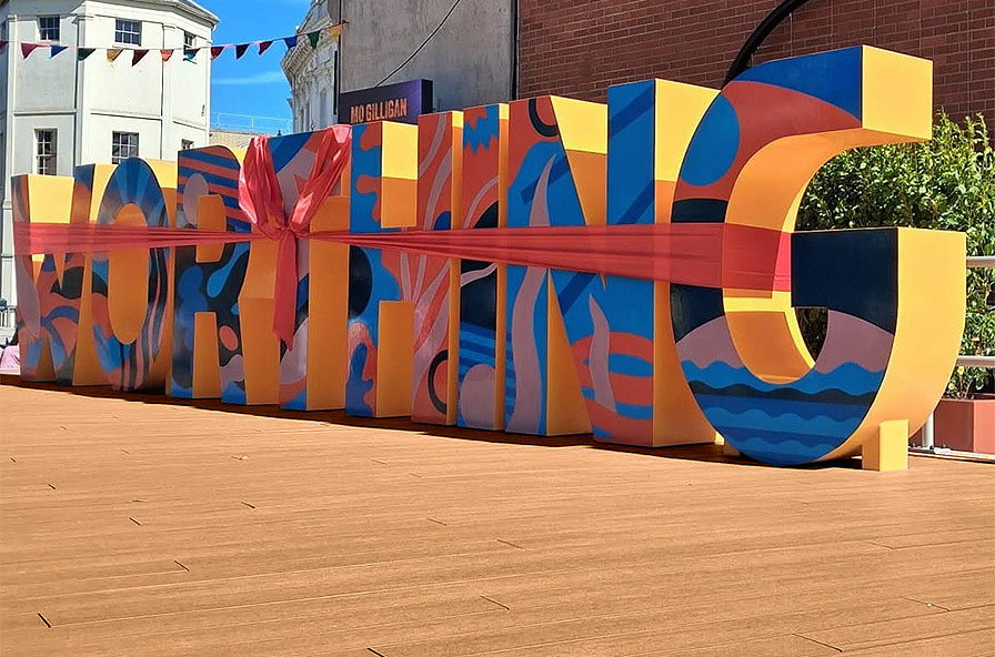 Large colourful sculpture spelling WORTHING on composite decking with a red ribbon around it