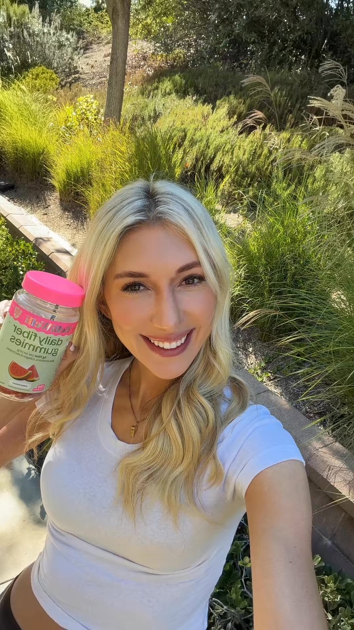 Smiling blonde woman in a white tee holding up a jar of BelliWelli Watermelon daily fiber gummies in a sunny garden