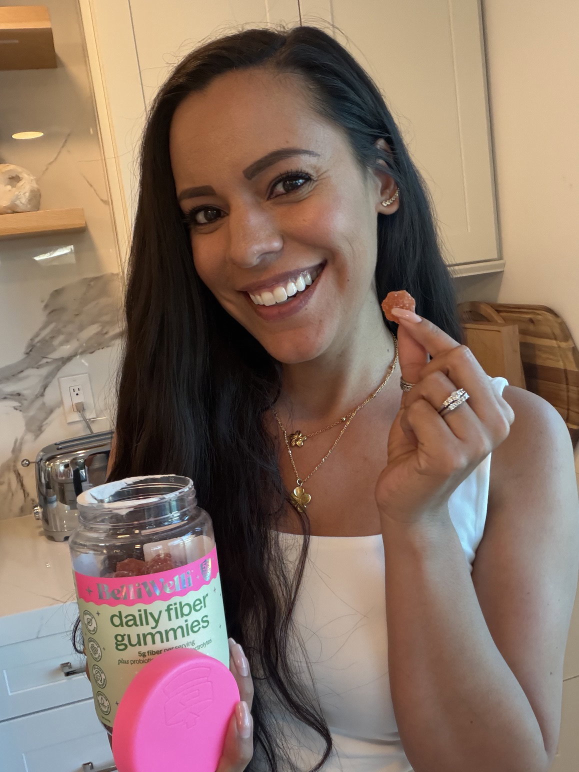 Smiling woman in her kitchen holding up a gummy and an open jar of BelliWelli watermelon daily fiber gummies