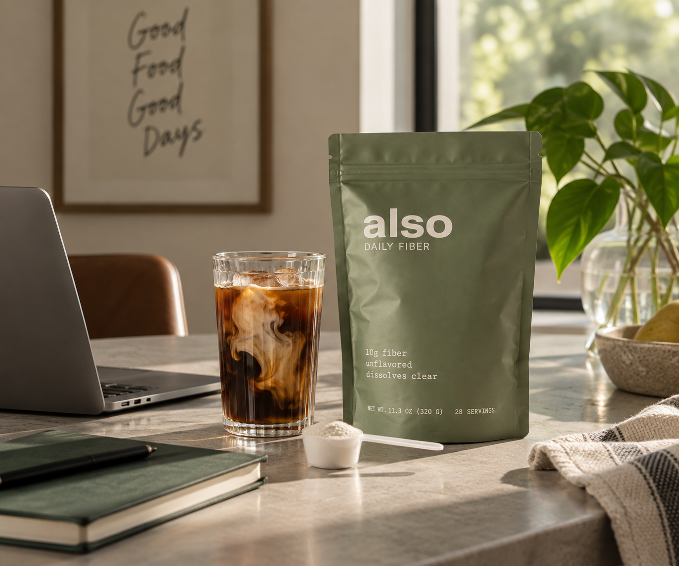 A pouch of ALSO Daily Fiber beside a glass of iced coffee and a scoop of fiber powder on a sunlit desk
