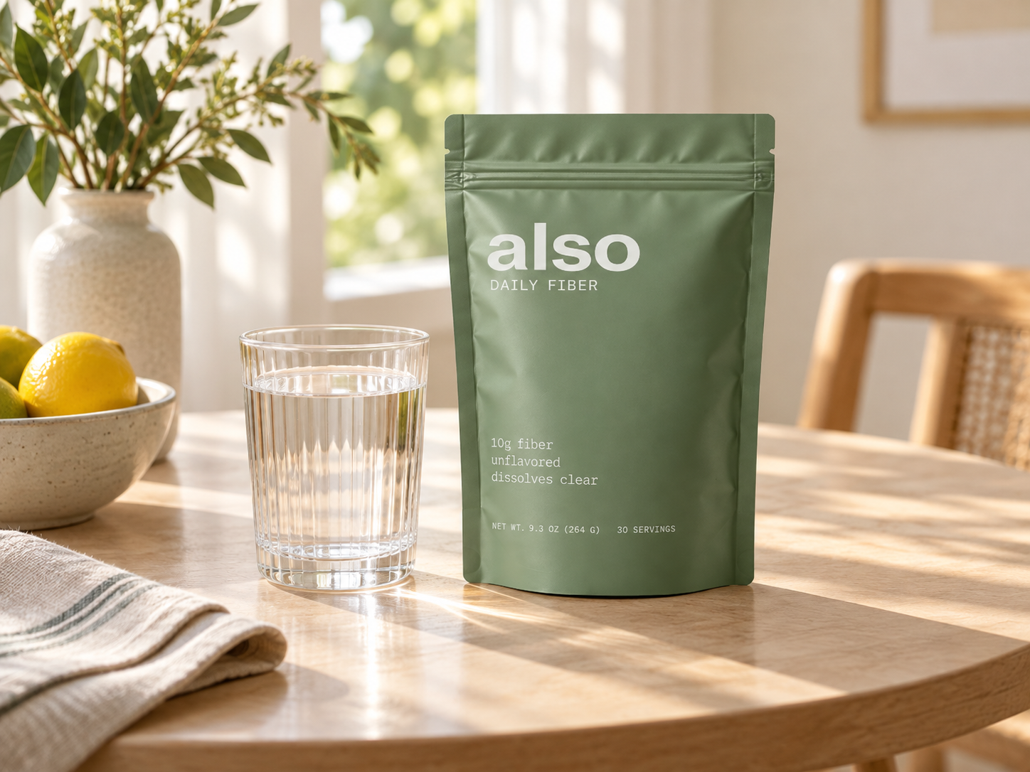 A pouch of ALSO Daily Fiber next to a glass of water on a sunlit wooden table