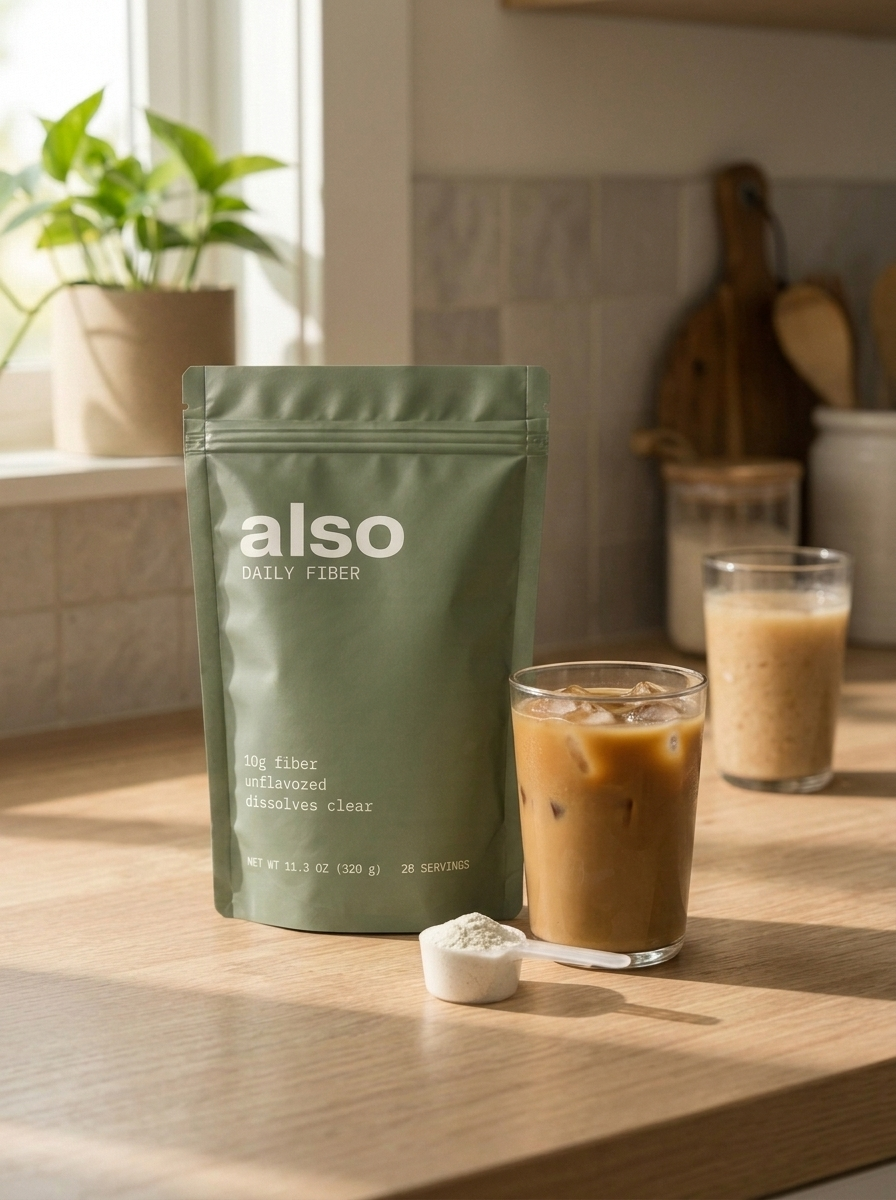 A sage green pouch of Also Daily Fiber beside a glass of iced coffee and a scoop of fiber powder on a sunlit wooden kitchen counter, with a potted plant on the windowsill behind