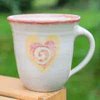 Keramikbecher mit Herzmotiv, rosafarbener Rand, auf Holzoberfläche, verschwommener grüner Hintergrund.