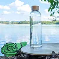 Klare Wasserflasche und grünes Handtuch auf einer runden Matte an einem See.