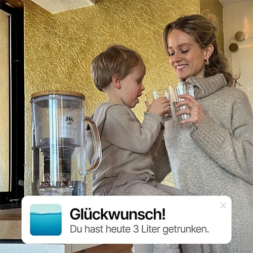 Frau und Kind in gemütlichem Zimmer, lächelnd mit Wassergläsern, Wasserfilter in der Nähe.