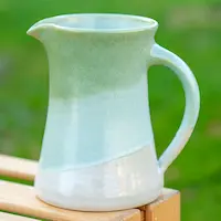 Keramikkrug mit grüner und weißer Glasur auf einer hölzernen Oberfläche, verschwommener grüner Hintergrund.