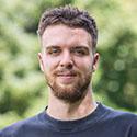 Mann mit kurzen Haaren und Bart lächelnd im Freien mit unscharfem grünen Hintergrund.