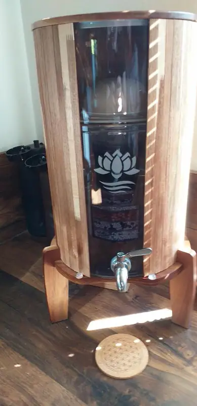 Wasserspender aus Holz von Lotus Vita, Metallzapfhahn und rundem Untersetzer darunter.