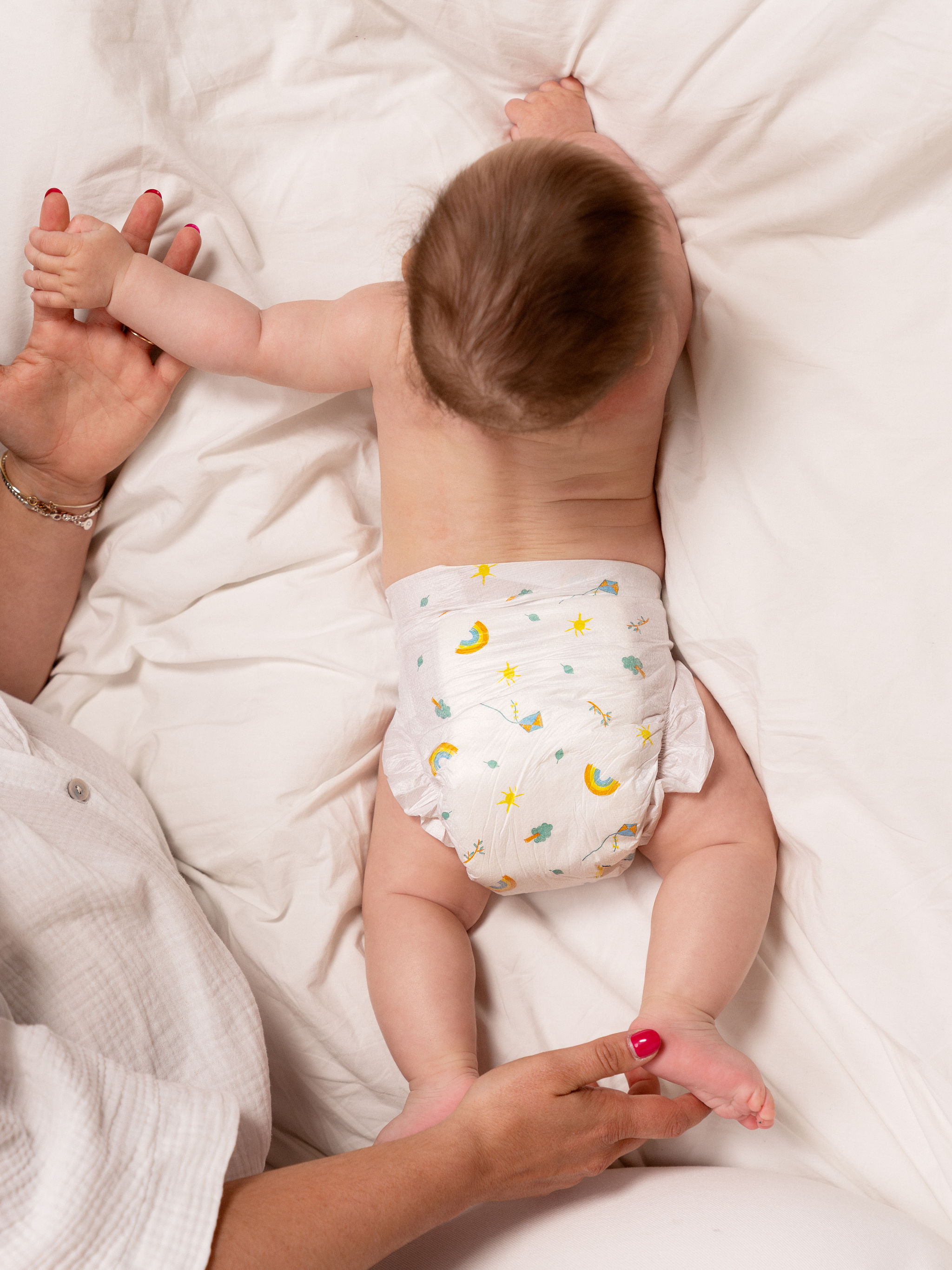Vue de dessus d'un bébé allongé sur un lit blanc portant une couche écologique à motifs, les mains de sa maman tenant les siennes