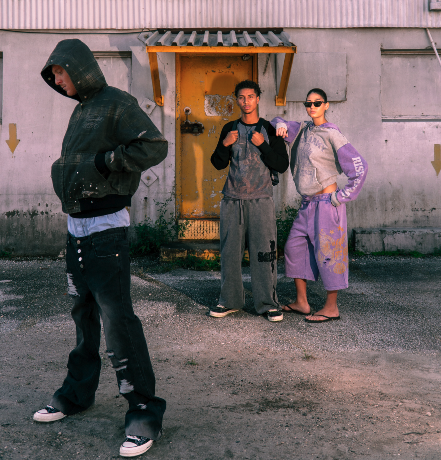 Three models in riseandfall Reconstruction pieces: a distressed plaid hooded jacket, a mesh raglan tee with grey sweatpants, and a lavender Rise Army hoodie with shorts, in front of a weathered yellow door