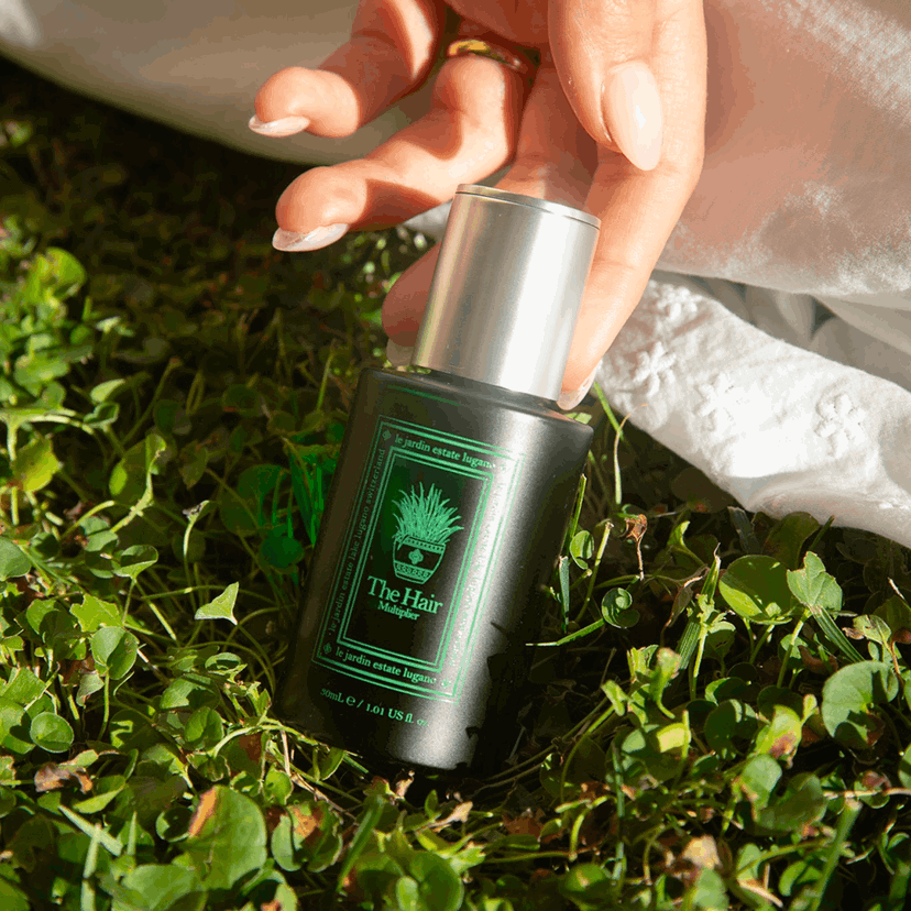 A hand reaching for The Hair Multiplier bottle resting on clover in sunlight