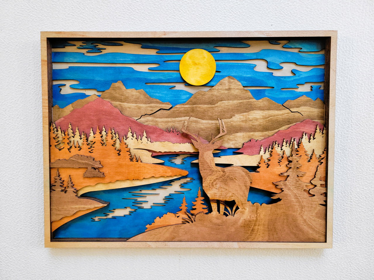 Layered wooden artwork of a deer by a river with mountains and a sun in the background