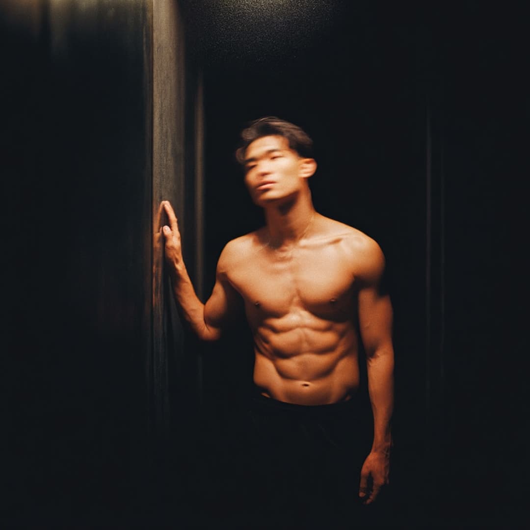 Blurry shirtless man stands by a dark wall, showcasing toned physique.