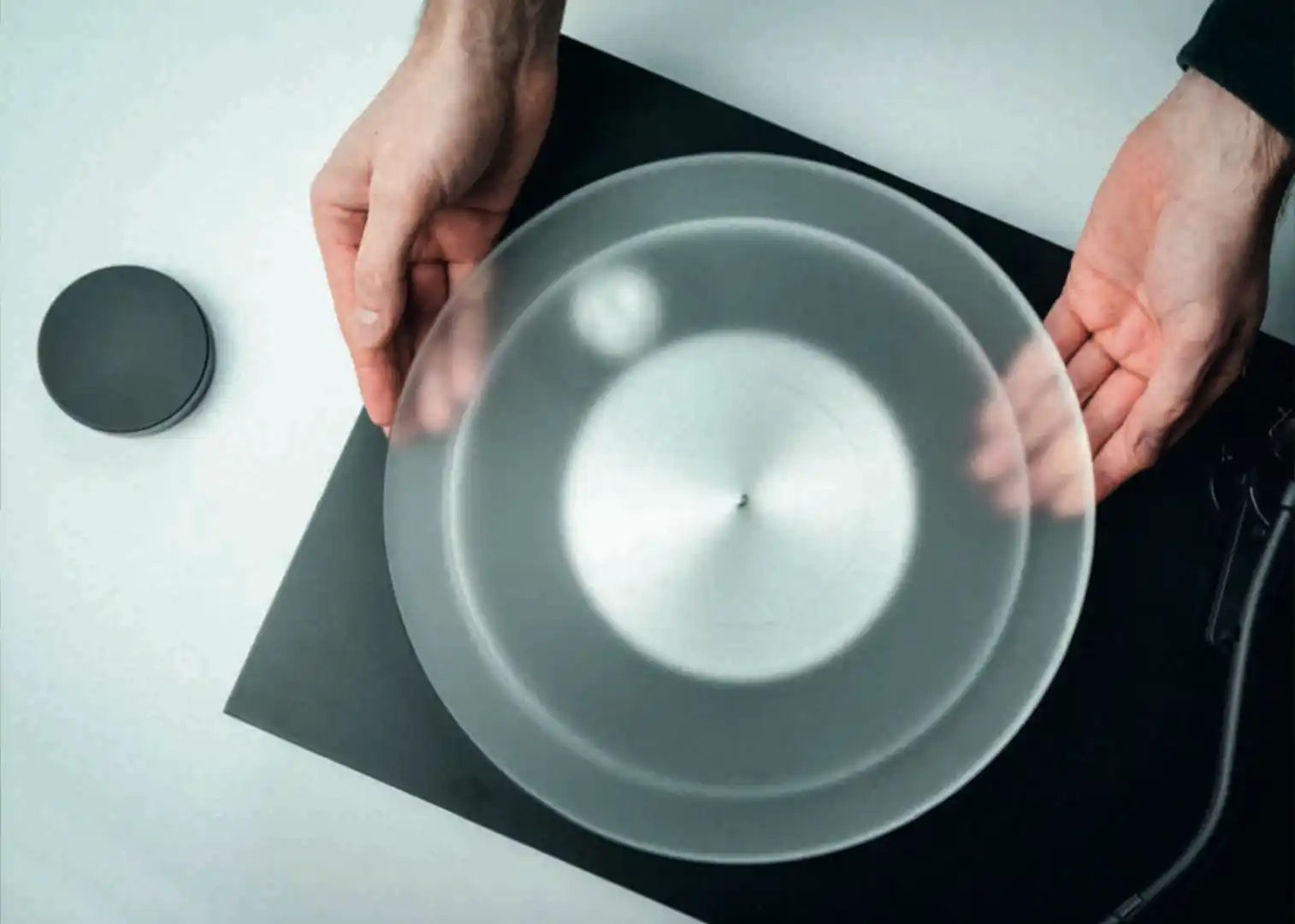 Two hands lowering a frosted acrylic platter onto a turntable spindle, with a dark slipmat resting on the table alongside