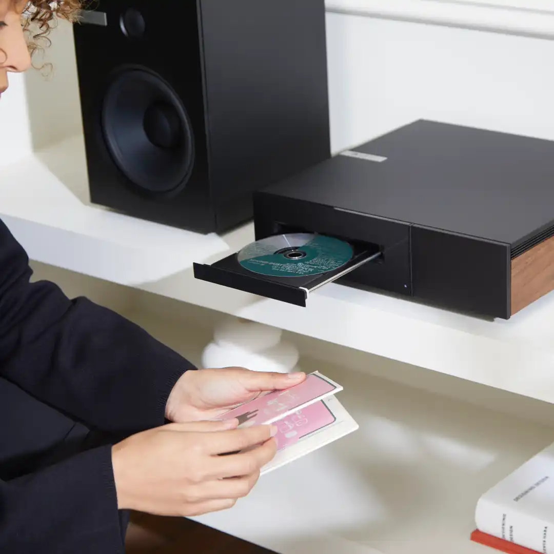 A listener loading a disc into the open tray of a black Cambridge Audio Evo CD transport with walnut side panels, on a white shelf beside a black bookshelf speaker