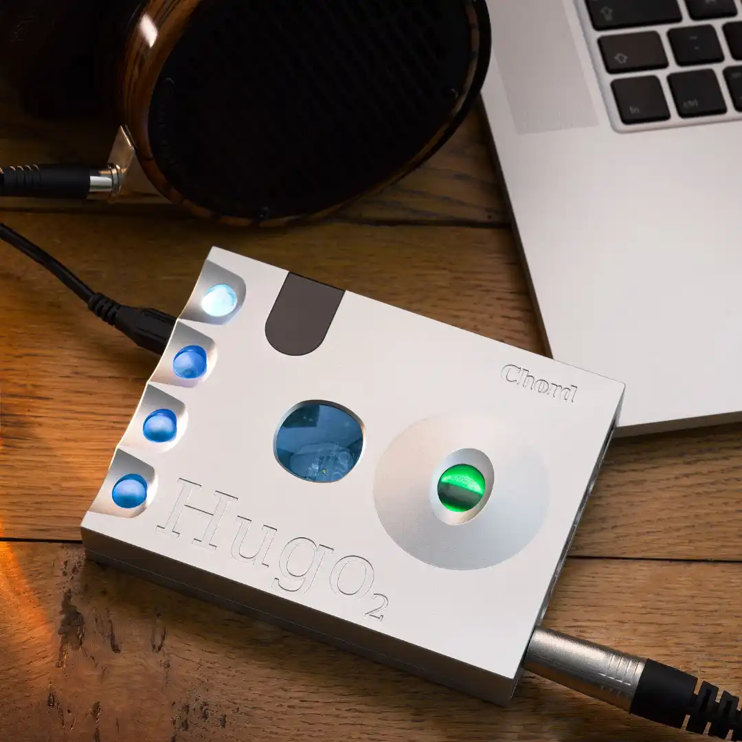 Silver Chord DAC headphone amplifier with glowing blue and green control orbs on a wooden desk, beside a laptop and wood-cup headphones