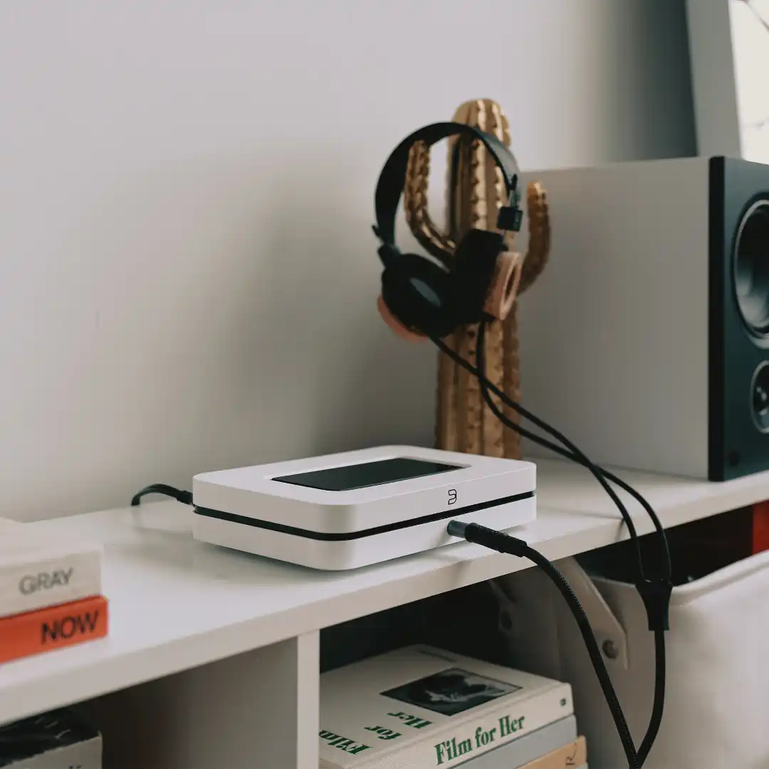 A black Bluesound Node music streamer on a walnut credenza in a dim living room, lit by a desk lamp, with a smartphone beside it