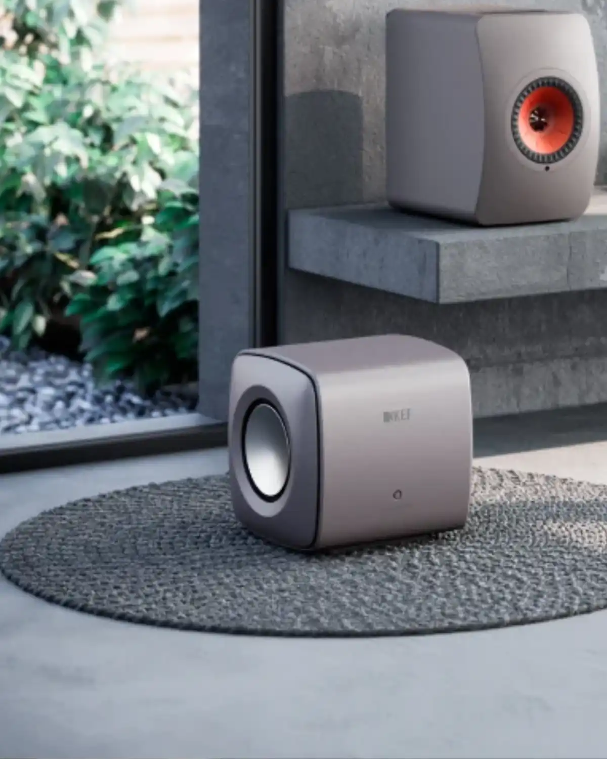 Compact KEF KC62 subwoofer in titanium grey on a round rug, with a matching KEF LS50 speaker on a stone shelf behind it
