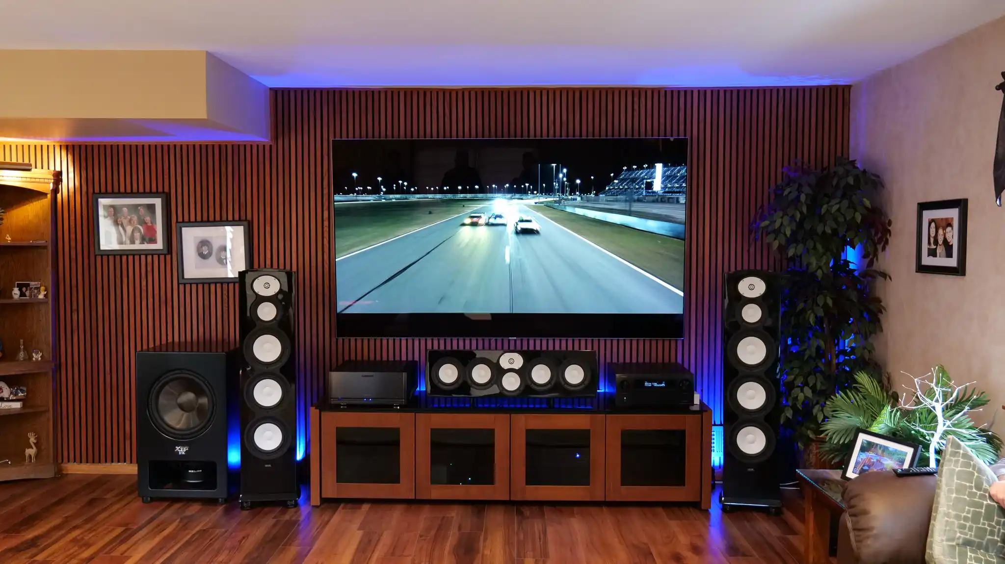 Media room with a large wall-mounted TV on a wood slat wall, gloss black floorstanding speakers, a center channel and a subwoofer