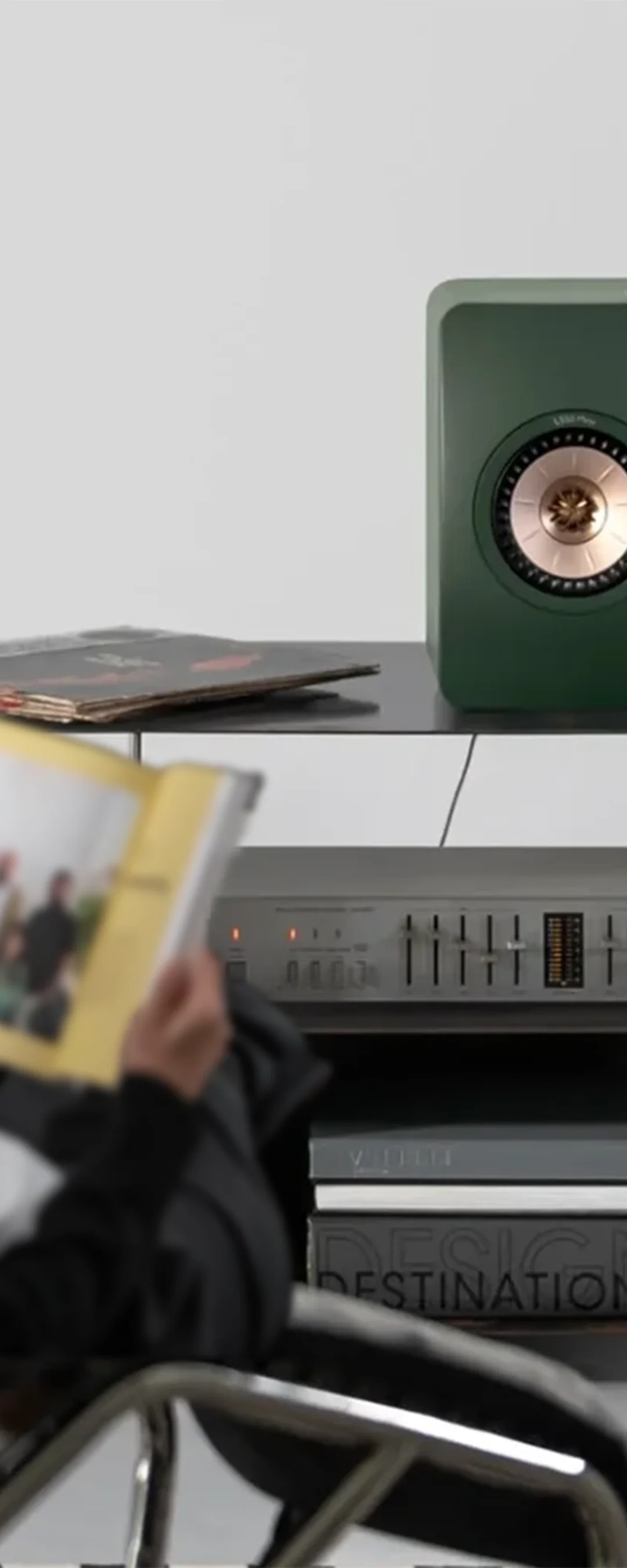 Listener reading a magazine beside a KEF bookshelf speaker and a hi-fi amplifier
