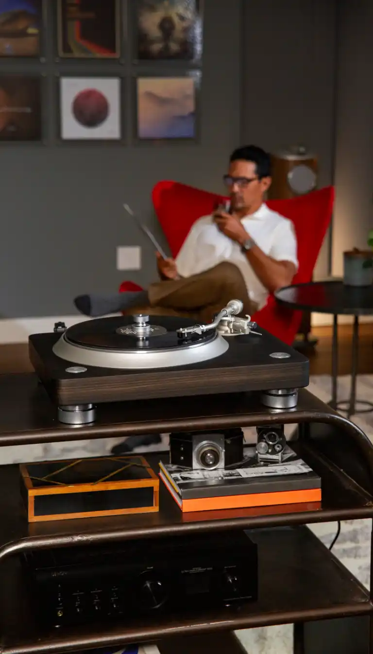 A woman lowering the tonearm onto a record on a black turntable, set on a rattan-front record cabinet next to a walnut floor-standing speaker