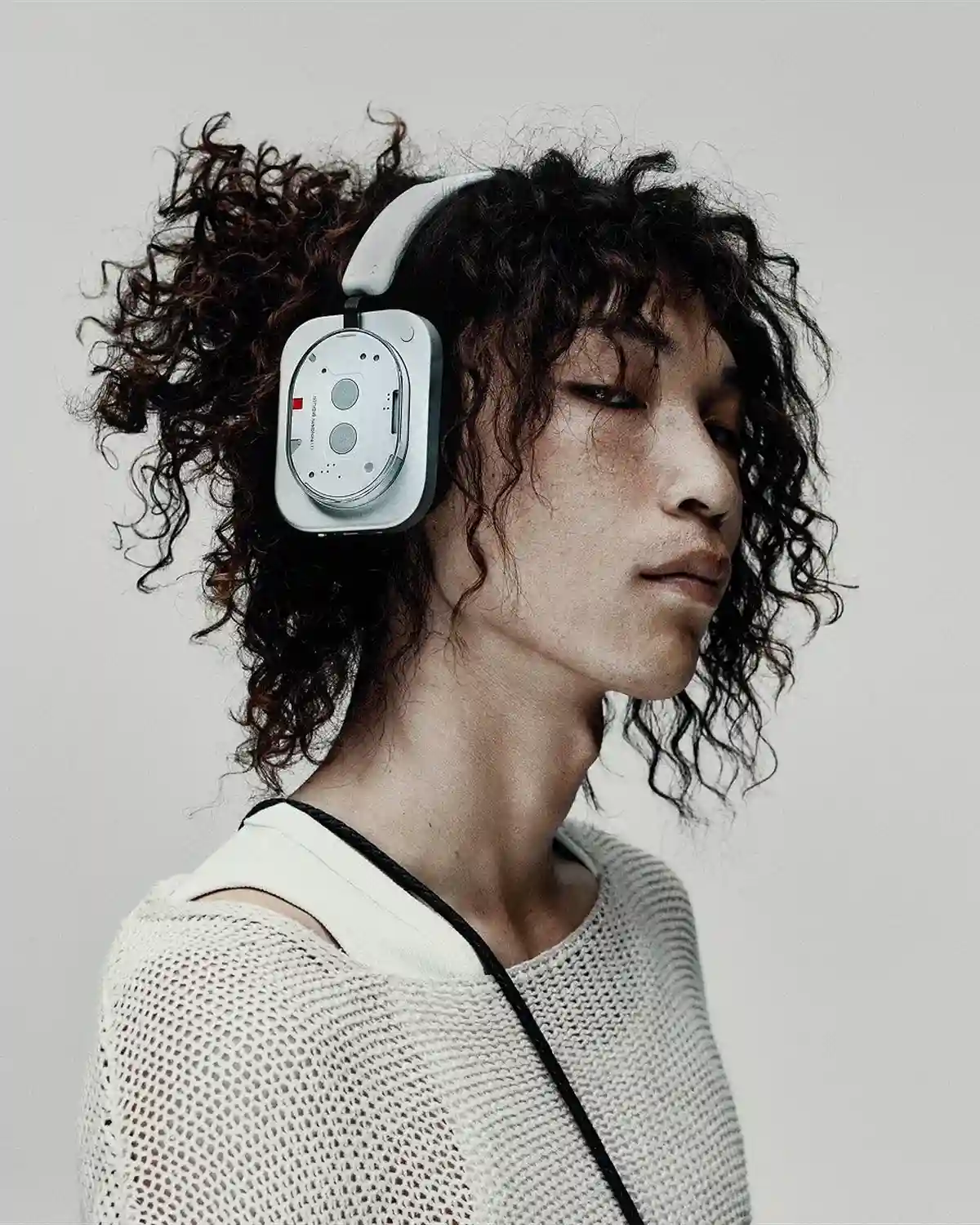 Person with curly hair wearing white Nothing Headphone (1) over-ear headphones, with sound by KEF, against a pale grey backdrop