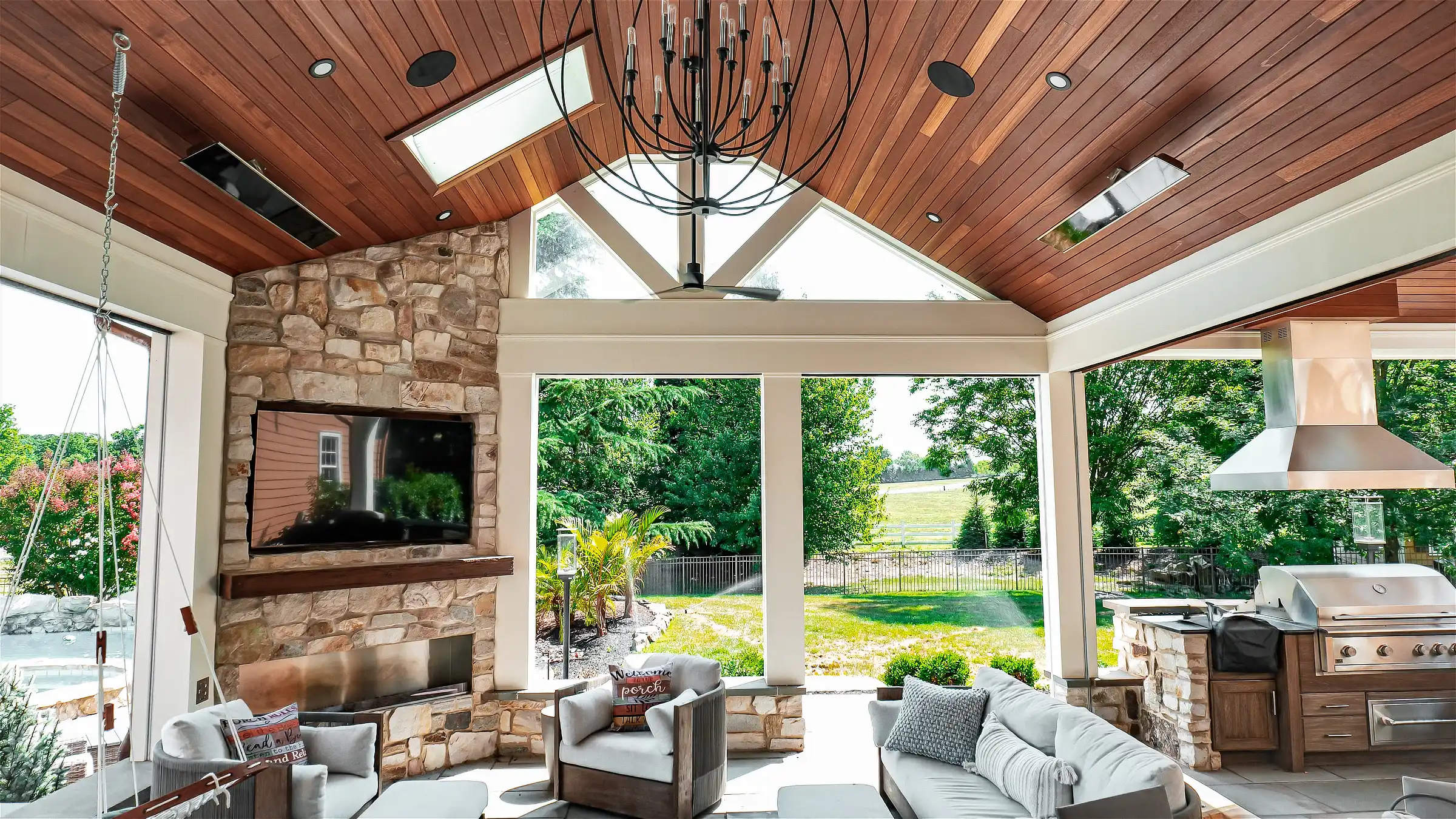 Covered outdoor living room with a stone fireplace TV, ceiling speakers, lounge seating and an outdoor kitchen