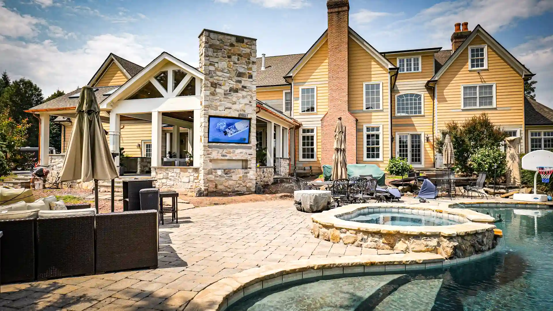 Backyard of a custom home with an outdoor TV built into a stone chimney beside a covered pavilion, overlooking the pool and spa