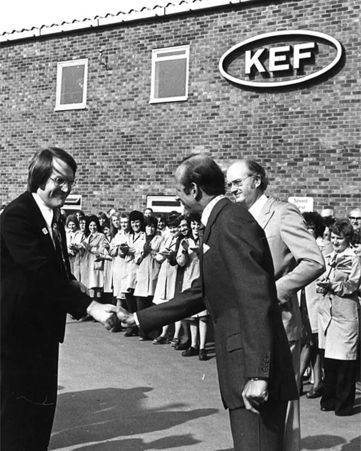 Archive black-and-white photo of a handshake outside the KEF factory in Maidstone as lines of staff applaud