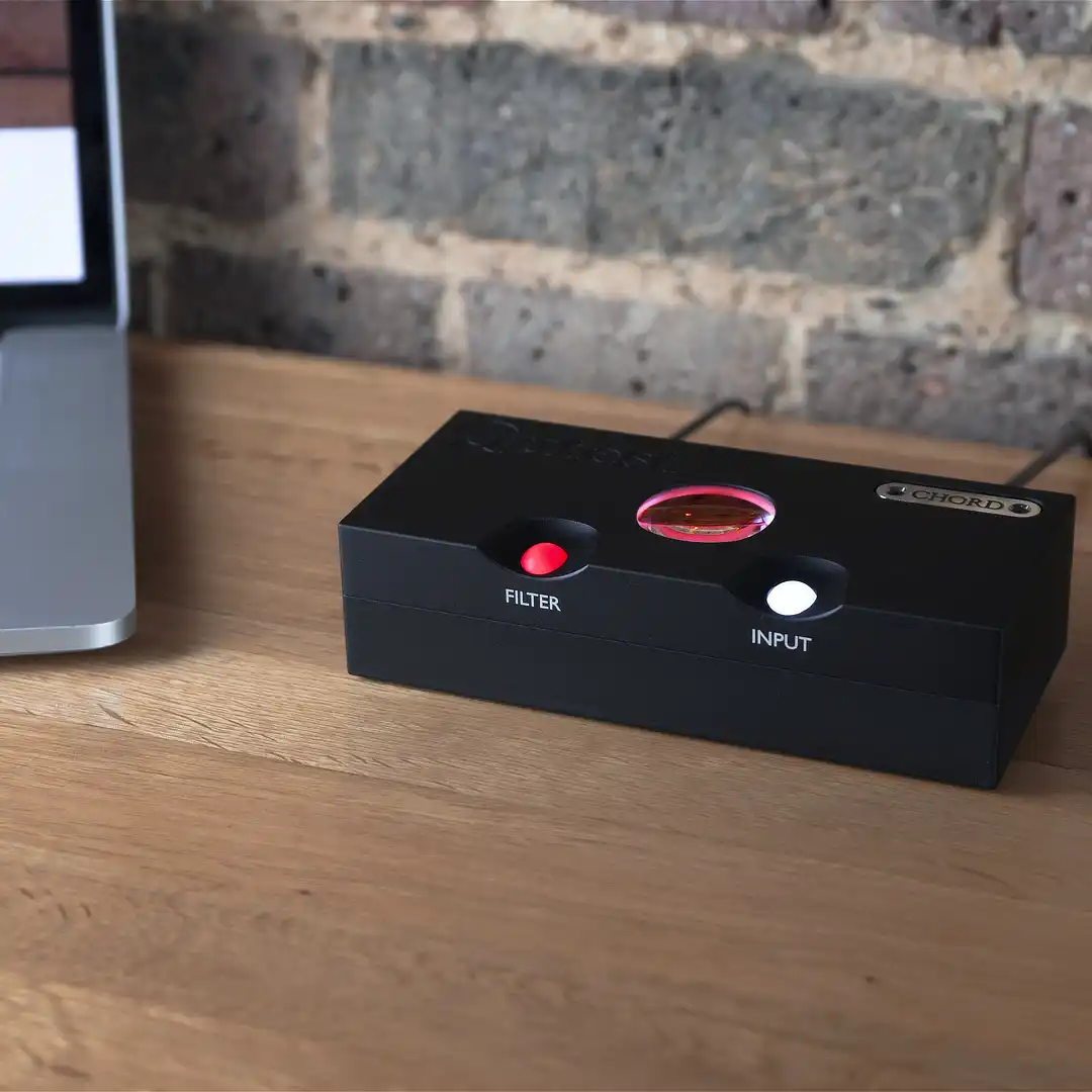 Black Chord Qutest DAC with its glowing filter and input buttons on an oak desk beside a laptop, in front of a brick wall