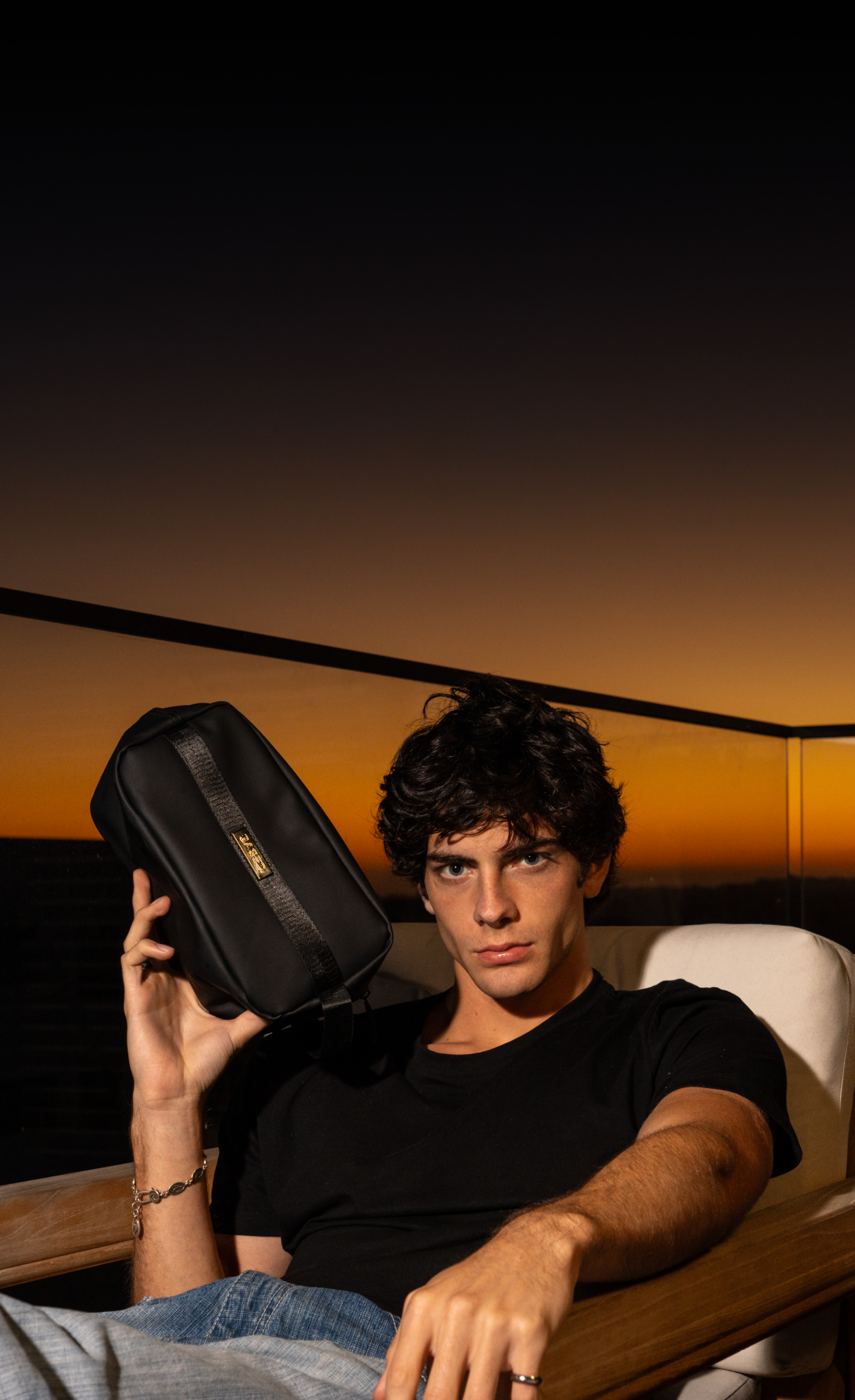 A young man in a black t-shirt holds a black bag, sitting on a chair with a sunset backdrop.