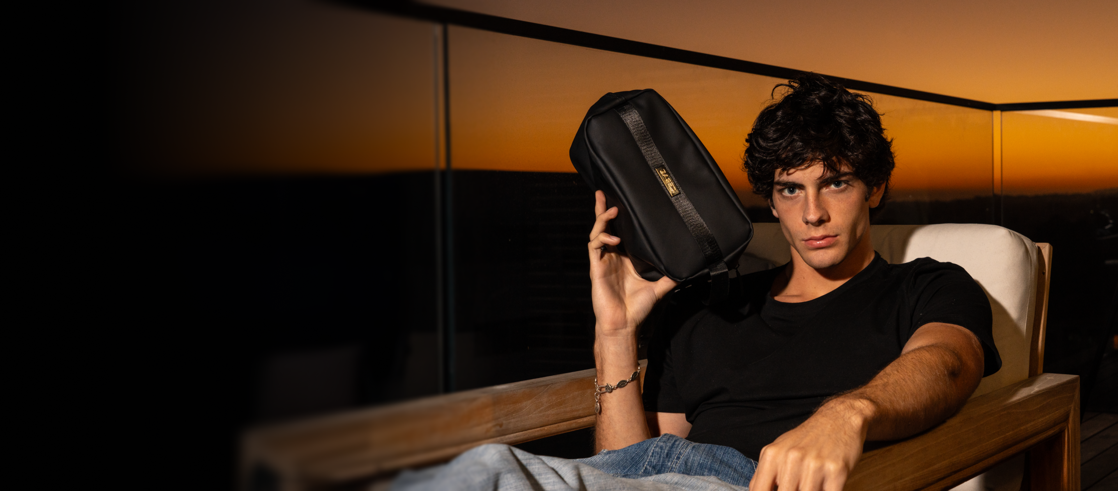 A young man in a black t-shirt holds a black bag while sitting on a wooden chair, with a sunset backdrop.