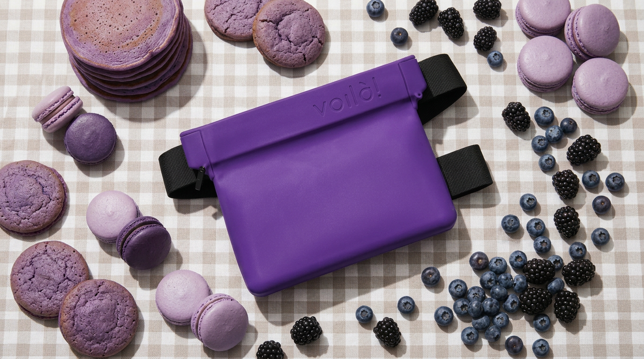 A purple toiletry bag surrounded by purple macarons, cookies, pancakes, and berries on a checked surface