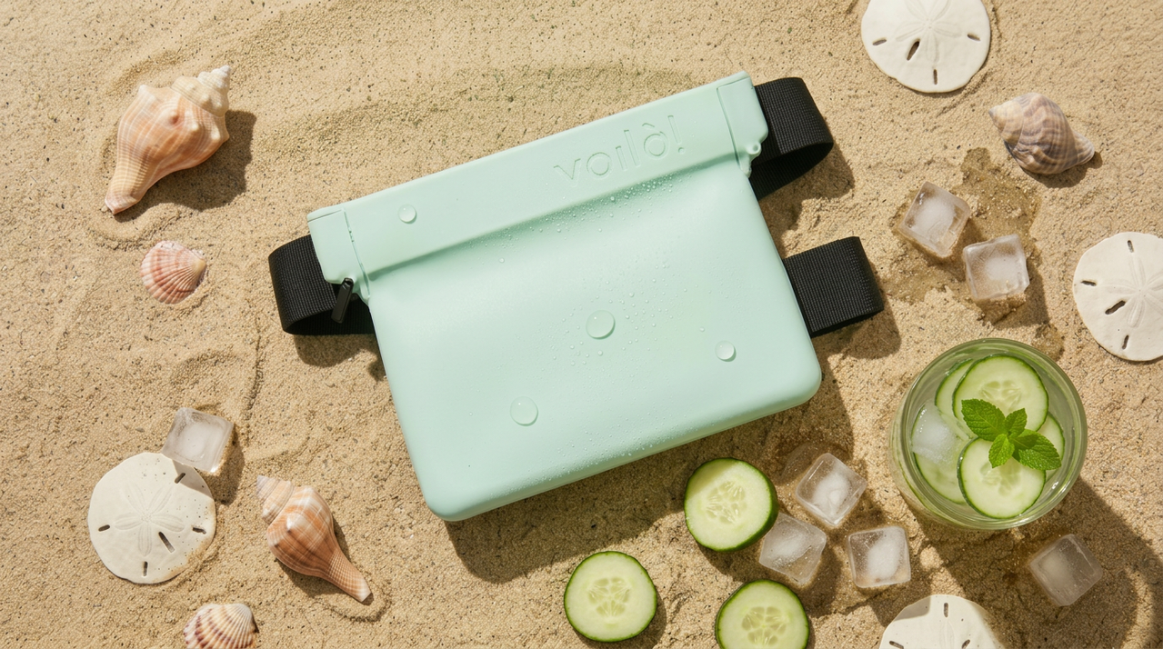 Mint green waterproof pouch on sandy beach surrounded by seashells, cucumber slices, ice cubes, and a cucumber drink