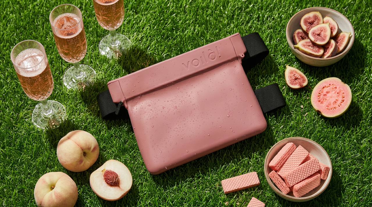 Pink waist bag on grass surrounded by pink wafers, sliced peaches, figs, and glasses of pink beverage