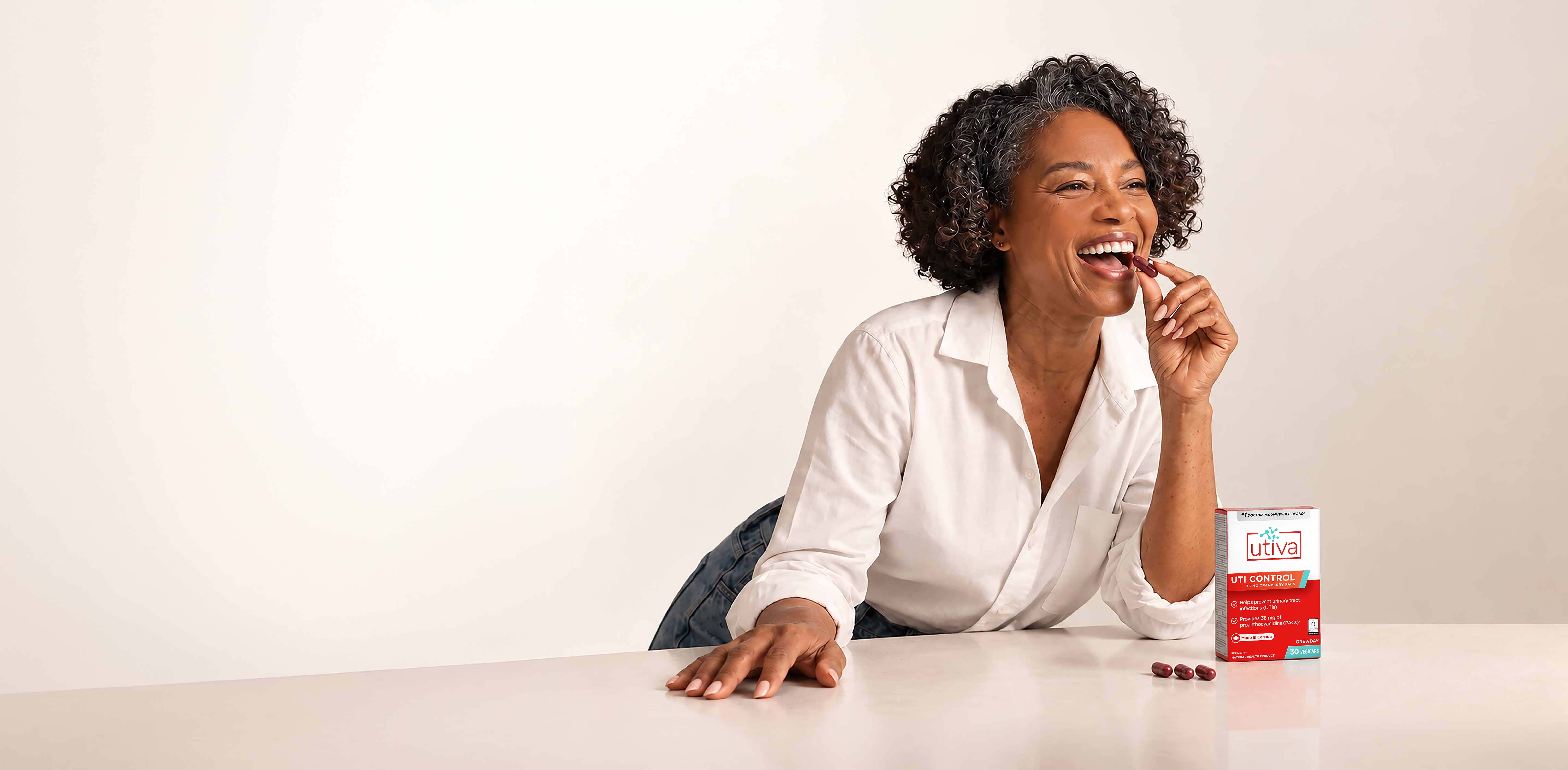 Smiling woman taking a Utiva UTI Control capsule at a table beside the product box