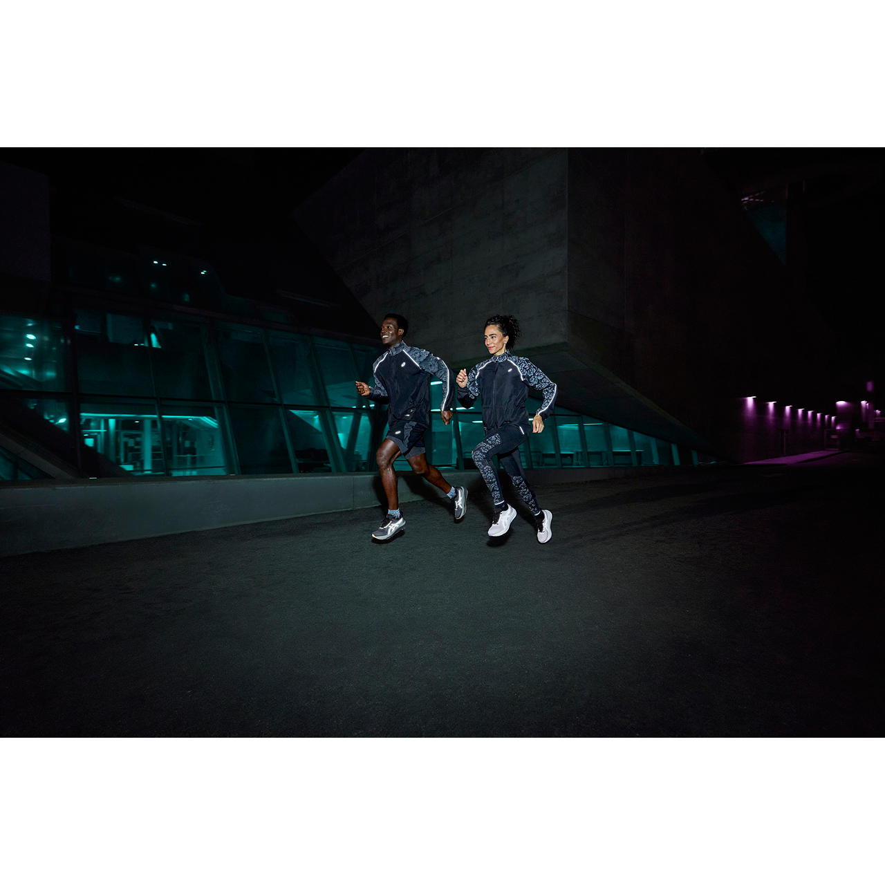 Two runners wearing ASICS Lite-Show reflective kit running past a lit glass building at night