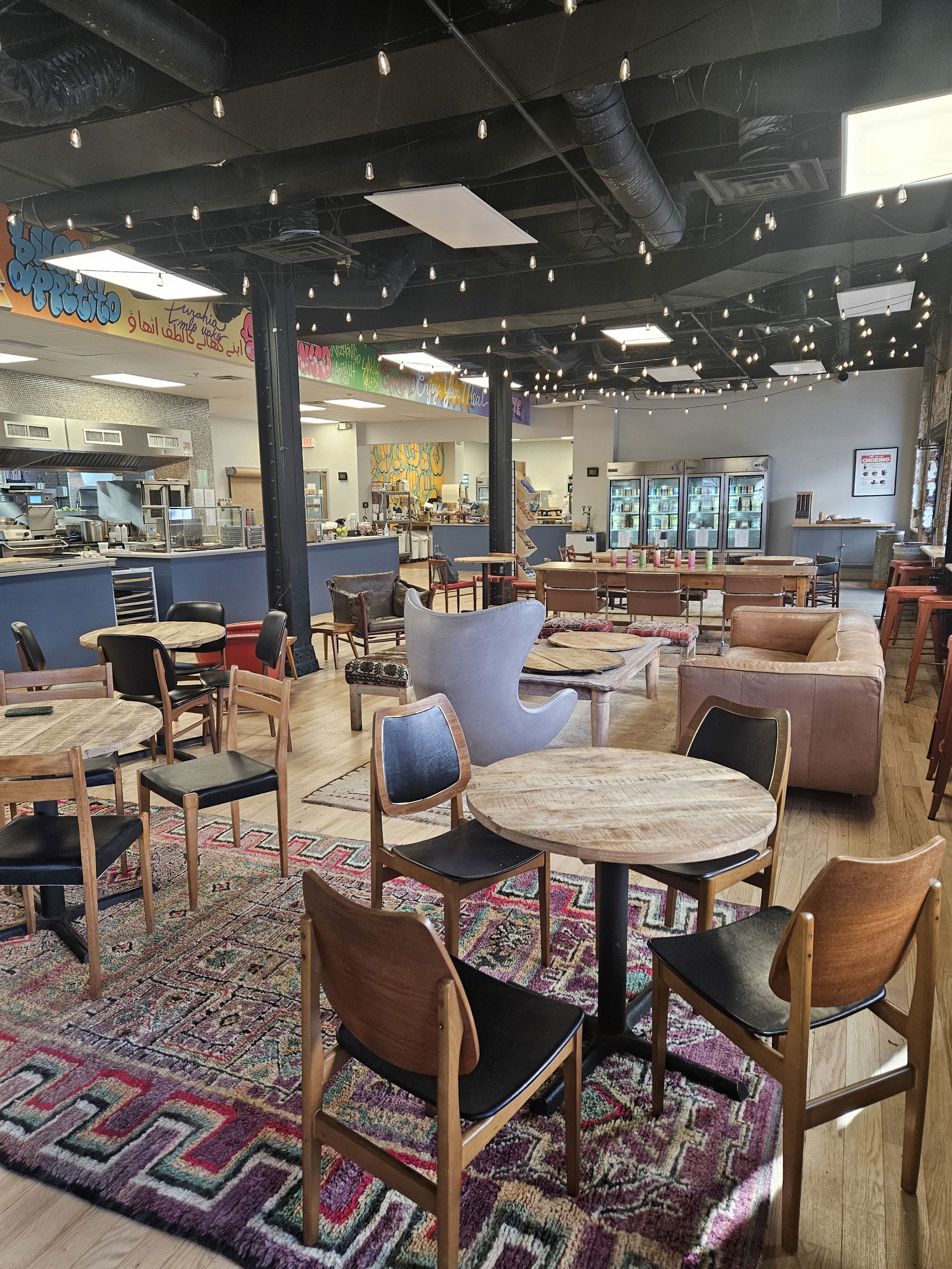 Cozy cafe with round tables, wooden chairs, colorful rugs, and string lights hanging from the ceiling.