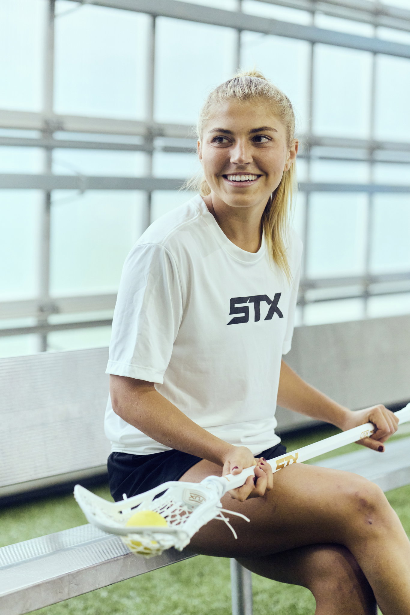 Chloe Humphrey sitting on a bench in an STX tee, smiling, her white Crux Pro across her lap with a yellow ball in the pocket