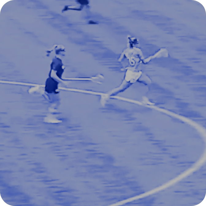 Two female lacrosse players running on a field, one in white and the other in dark uniform.