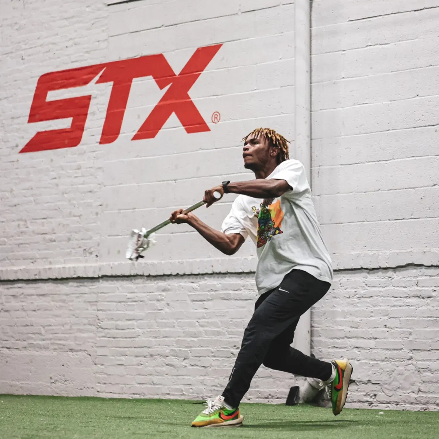 Person playing lacrosse indoors, wearing casual attire. Large STX logo on the white brick wall behind.