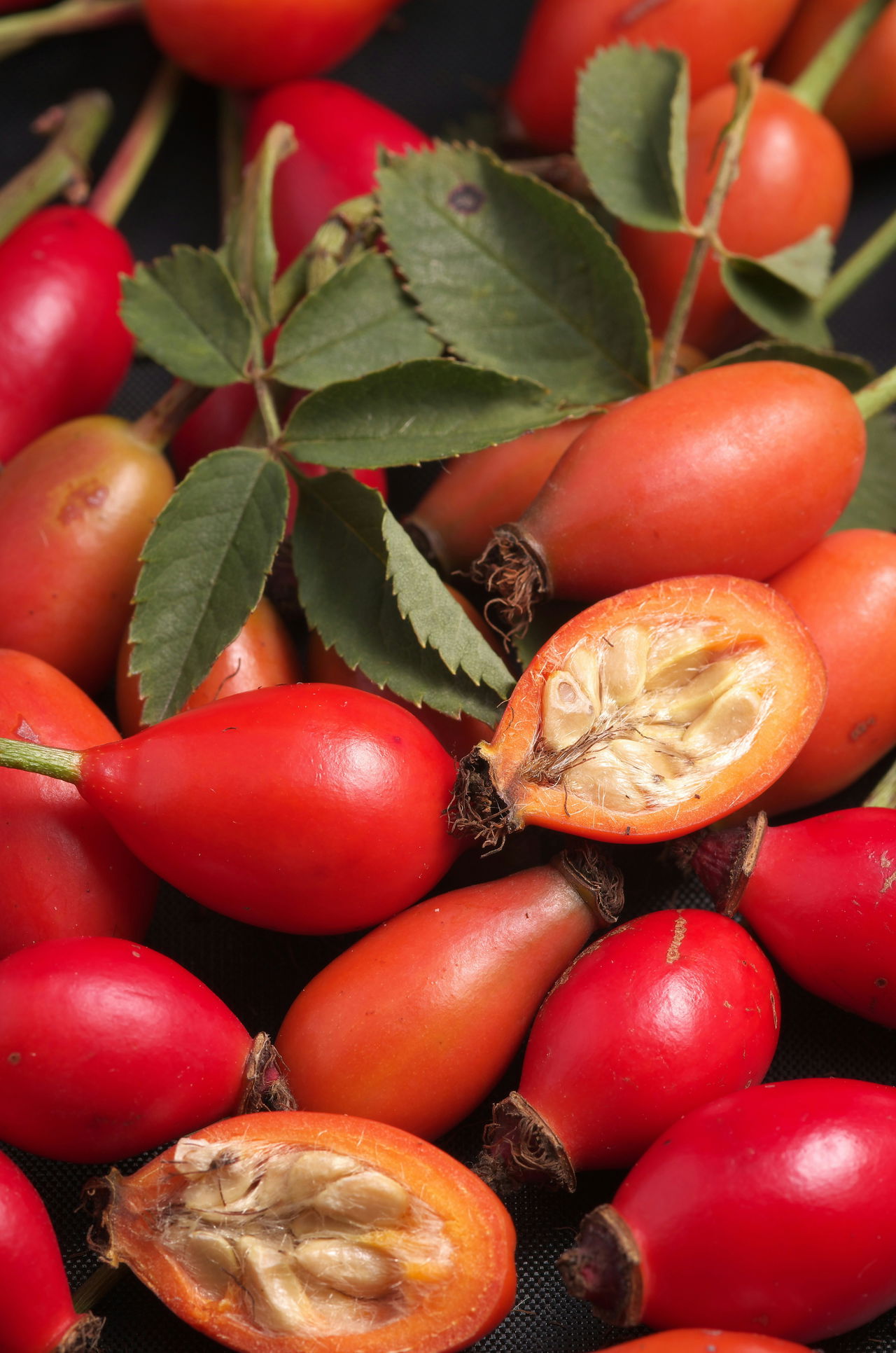 Close-up of red and orange rose hips with green leaves, one cut open showing seeds inside.