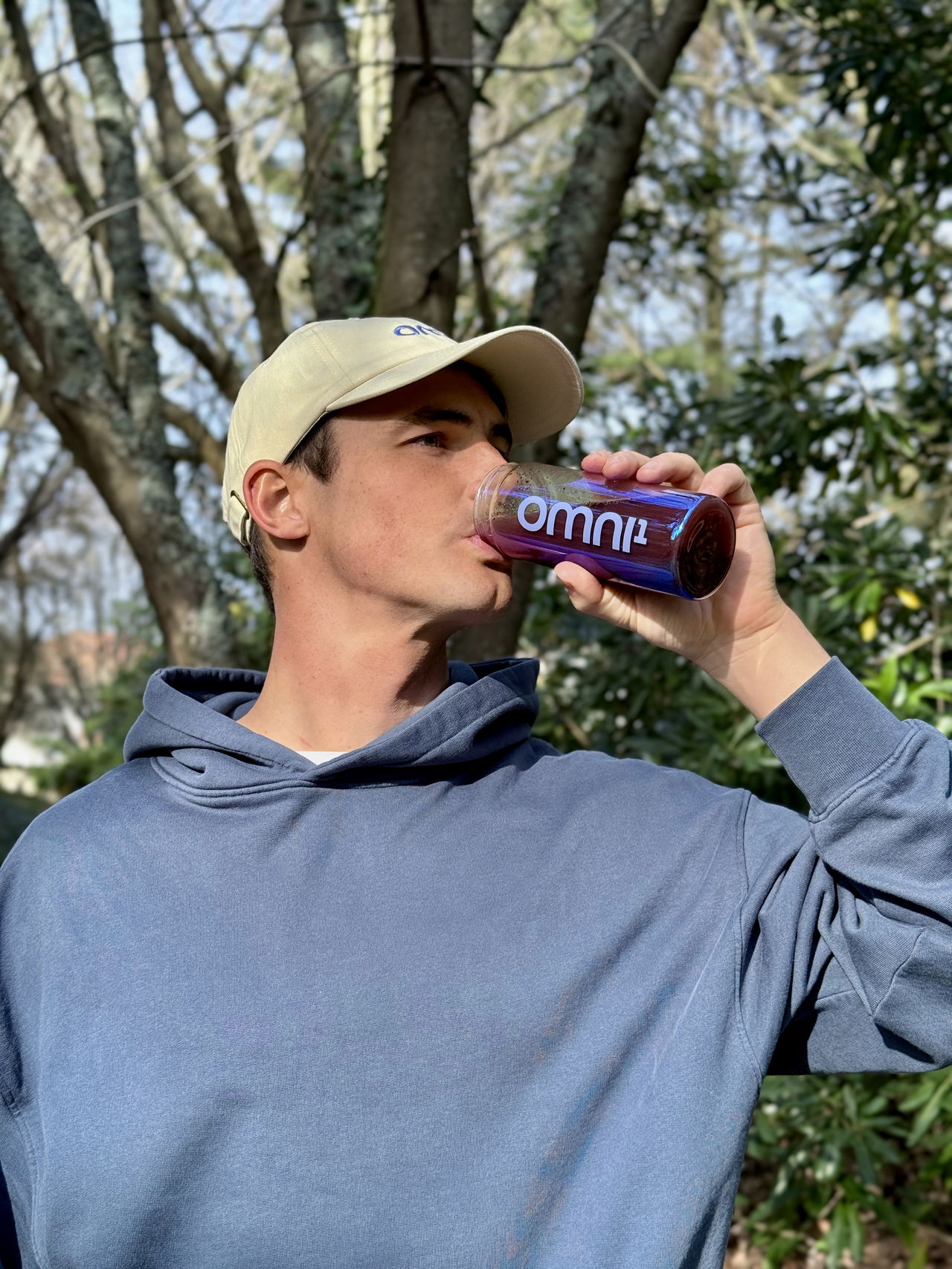 Man in cap and hoodie drinking a purple drink from a bottle labeled OMNI¹ in a wooded area