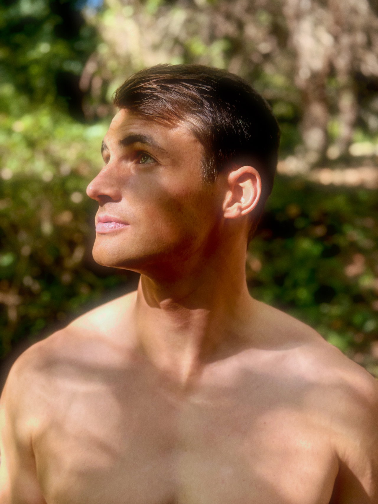 Shirtless man with short hair looking up with dappled sunlight on his face and shoulders