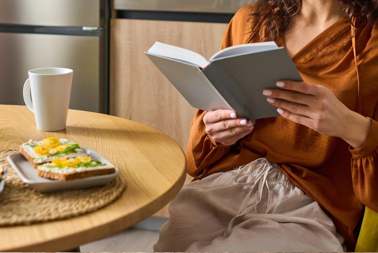 Donna seduta in cucina mentre legge un libro e fa colazione: un momento di relax dopo aver seguito i consigli per le pulizie estive di Mamma Avilia.