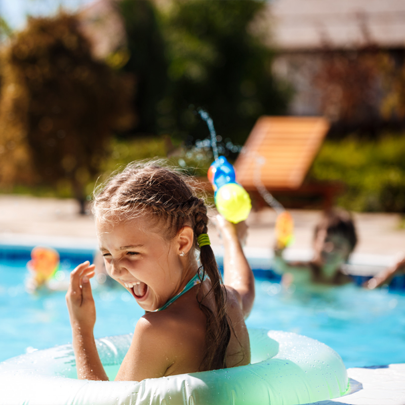 Bambina che gioca e ride in piscina con un salvagente e pistola ad acqua: l’estate perfetta per i più piccoli secondo i consigli di Mamma Avilia.