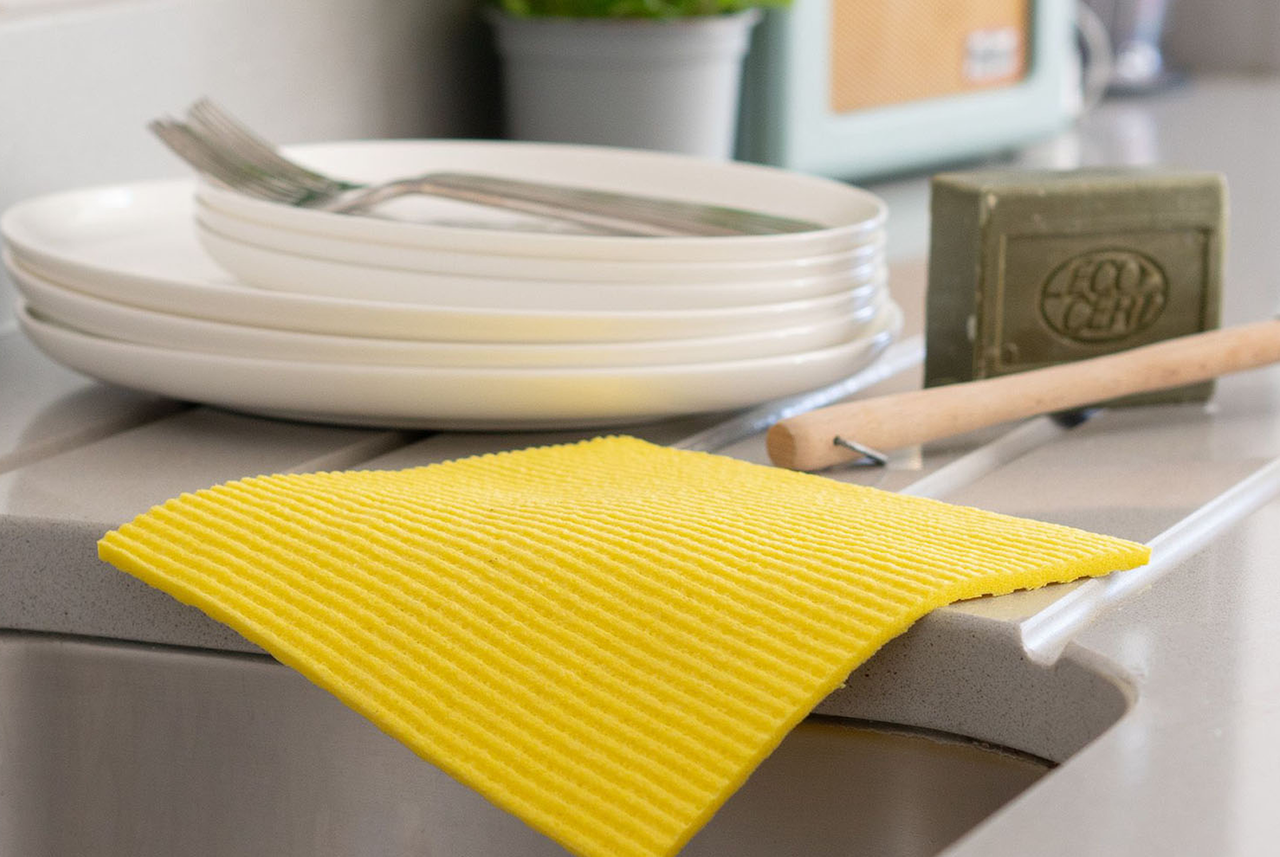 Panno giallo da cucina accanto a piatti puliti, posate e sapone naturale: i consigli di Mamma Avilia per igienizzare superfici e stracci in modo semplice e sicuro.