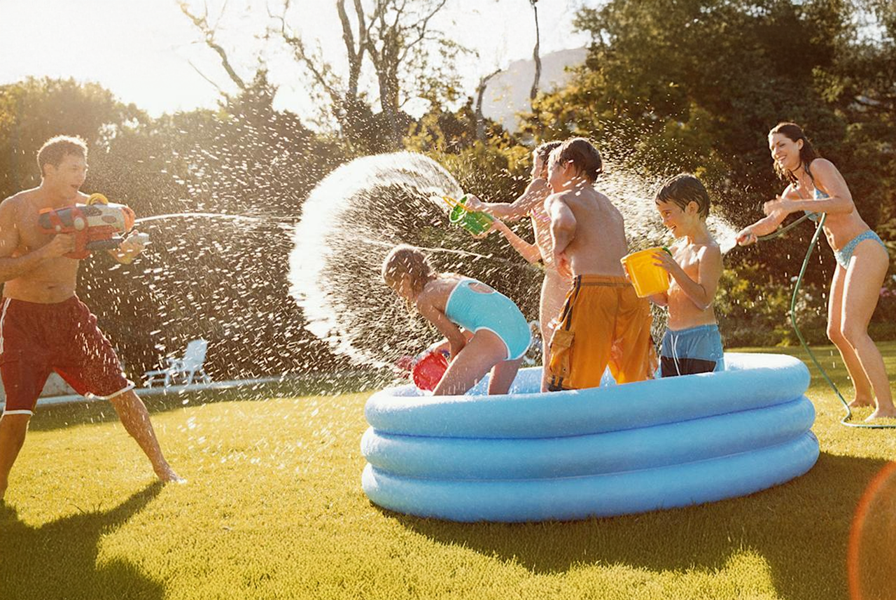 Bambini e genitori che giocano con pistole ad acqua e secchi in giardino intorno a una piscina gonfiabile: l’estate all’aria aperta secondo Mamma Avilia, tra risate e giochi d’acqua.